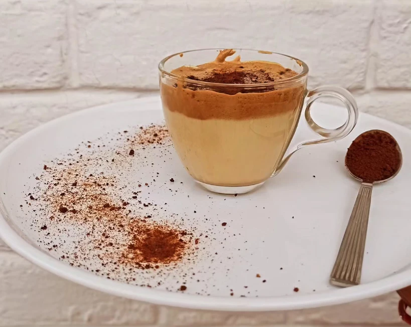 Glass of Dalgona coffee on white plate sprinkled with instant coffee